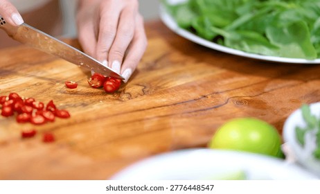 A person uses a sharp knife to slice spicy red chili peppers on wood.