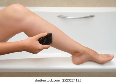 Person using electric epilator on leg while sitting on bathtub edge. Close-up photography for skincare and personal grooming concept - Powered by Shutterstock