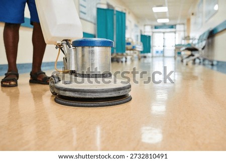 A person is using a floor buffer to clean and shine the polished floor in a hospital corridor with medical beds and curtains visible in the background
