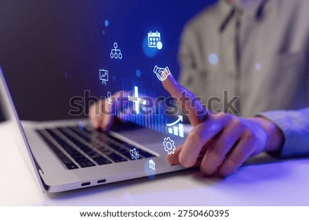 A person is using a laptop while touching a digital screen filled with icons. This scene takes place in the evening. The setting looks modern and focused on technology.