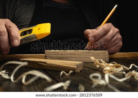 A person is using a measuring tool and a pencil to mark wood in a workshop. Wood shavings are scattered on the surface. The lighting is low, creating a focused work area.