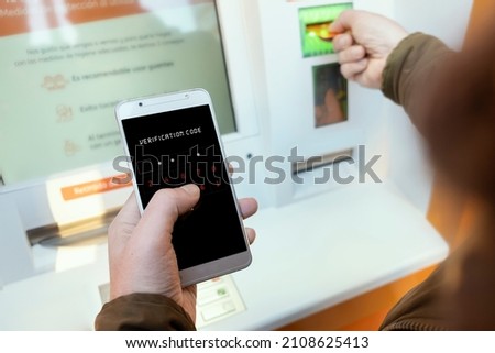 Person using mobile phone with verification code at ATM. Security concept for telephone authentication. Man using ATM with card and checking security code with mobile. Mobile to verify bank code