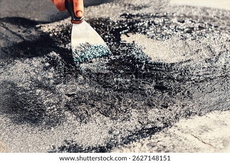 Person using a putty knife to spread a thick, black tar substance across a rough surface,  for roof repair. Material has a shiny, wet appearance with small reflective particles