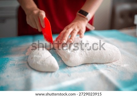 A person using a red bench scraper to divide dough on a floured blue surface in a baking process