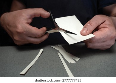 A person is using scissors to cut a piece of paper into strips at a work surface. The surrounding area has small paper scraps spread out. It is a moment of focused crafting.