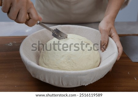 A person is using a scoring tool to make cuts on the top of a dough loaf in a banneton. The dough is risen and ready for baking.