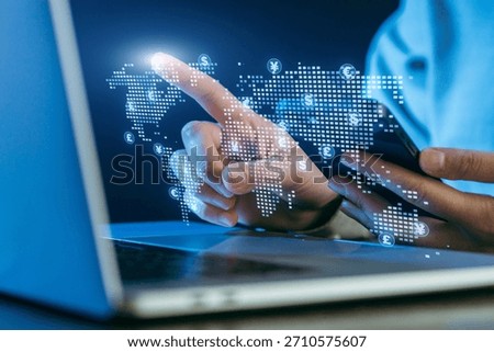 A person is using a smartphone while pointing at a digital map displayed on a laptop. The map shows currency symbols and represents global finance trends.