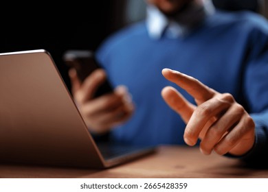 Person using smartphone while sitting at a wooden desk with a laptop in a dimly lit indoor setting