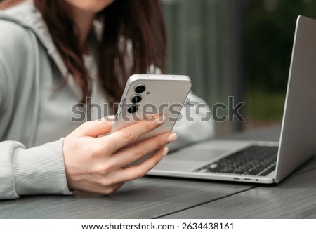 A person using a smartphone while sitting with a laptop on a wooden table outdoors, symbolizing multitasking and modern technology.