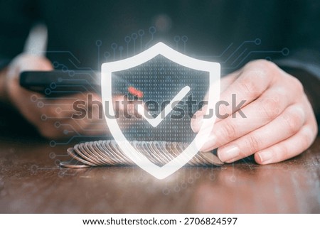 A person is using a smartphone while sitting at a table with coins. A shield icon appears with a check mark, symbolizing online security in finance and transactions.