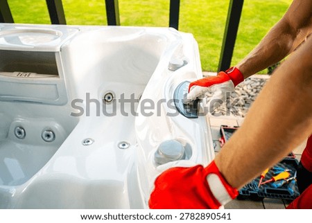 A person is using a tool to clean a hot tub at their home. The setting is bright and sunny with green grass visible in the background. Various cleaning tools are on the ground beside them.