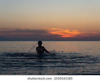 A person wades in the calm sea water during sunset, creating splashes as colors of orange and pink light up the sky. The peaceful setting evokes a sense of relaxation. - Powered by Shutterstock