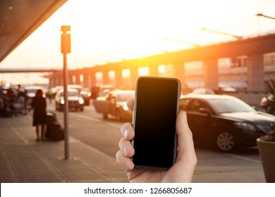 Person Waiting And Using Taxi App, Holding A Mobile Smartphone At An Airport Arrivals Taxi Line Or Ride Share Pick Up Location. Blank Screen. App Mockup