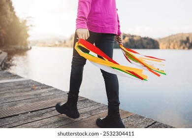 A person walking along a wooden dock by a lake, holding colorful ribbons in their hands. The scene is serene with trees and hills in the background, and the sunlight reflects on the water. - Powered by Shutterstock