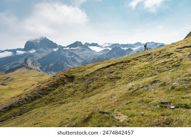 A person is walking up a grassy hill in the mountains under a cloudy sky, surrounded by lush greenery with a vast landscape spreading in the horizon. High quality photo - Powered by Shutterstock