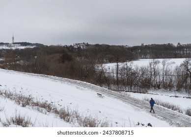 A person walks alone on a snowy path beside a frozen lake, with bare trees and a distant tower. - Powered by Shutterstock