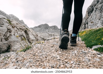 A person walks along a rocky path with hiking poles, flanked by rugged mountains and greenery under a cloudy sky. - Powered by Shutterstock