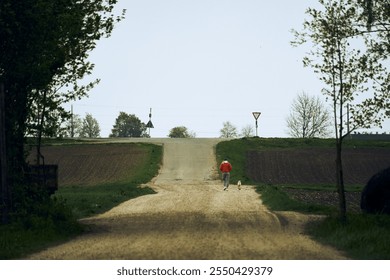 Person walks a dog along a quiet dirt road surrounded by fields and trees in rural landscape - Powered by Shutterstock