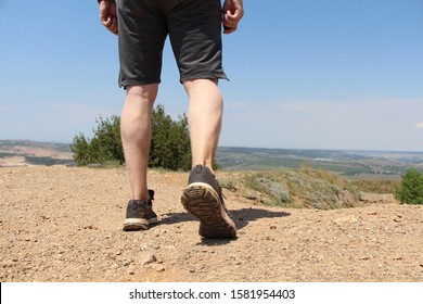 Person walks on the hill far away from the city. Close up view of legs in sport shoes. Man dressed in gray shorts. Sunny weather. Copy space for text.
