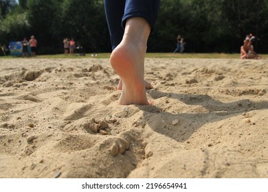 Person Walks On The Sand On The Beach With Bare Feet On Sunny Summer Day. Sand And A Foot Walking Away From The Camera Close-up. Some People Are Seen On The Background. Image With Selective Focus