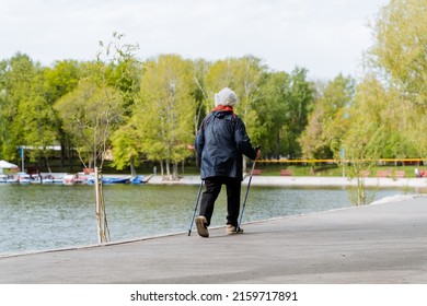 A Person Walks In The Park With Walking Poles, A Pensioner Is Engaged In Nordic Walking, A Summer Workout By The Lake, An Elderly Person. High Quality Photo