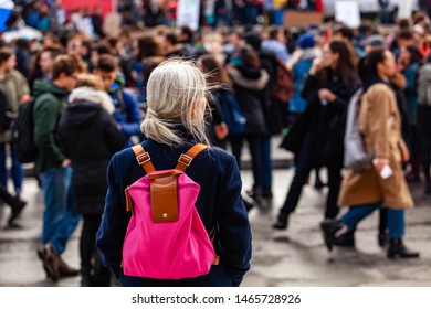Person watches ecological protest. An older woman is viewed from behind, wearing a pink shoulder bag, watching an environmental demonstration on a street in Montreal, Canada