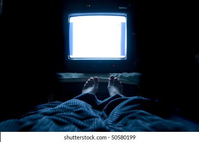 Person watches TV at night in his bed with his feet sticking up out of the blankets. The TV screen is blank white