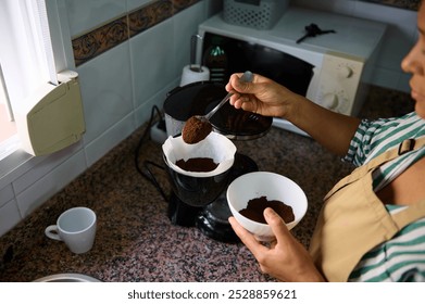 A person wearing an apron prepares coffee using a French press in a kitchen. Ground coffee is placed into the brewer, creating a cozy morning ritual. - Powered by Shutterstock