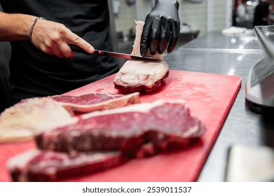 person wearing black gloves slices raw meat on red cutting board, with several steaks nearby. image highlights food preparation and culinary skills, suitable for cooking and kitchen-related themes. - Powered by Shutterstock