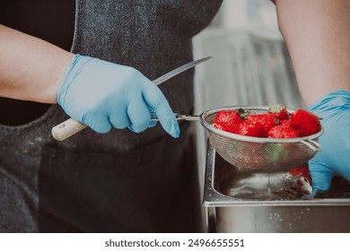 Person wearing blue gloves is washing strawberries in a metal colander in a professional kitchen. The strawberries are being washed in a stainless steel sink - Powered by Shutterstock