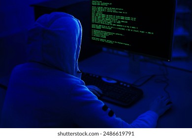A person wearing a hooded sweatshirt sits in front of a computer with a dark background. Computer screen displays green code, indicating that the person is working on a programming or technical task.