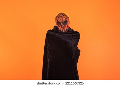 Person With Wearing Jack O Lantern Pumpkin Mask And Black Hooded Cape, Celebrating Halloween, On Orange Background. Celebration Concept, All Souls' Day And All Saints' Day.