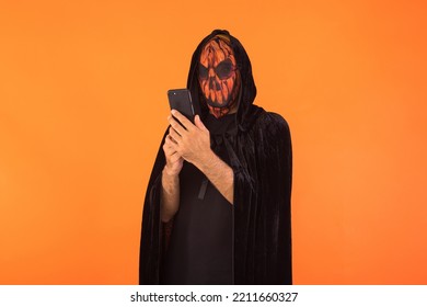 Person Wearing Jack O Lantern Pumpkin Mask And Black Hooded Cape, Consulting His Mobile Phone, Celebrating Halloween, On Orange Background. Celebration Concept, All Souls' Day And All Saints' Day.