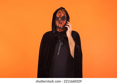 Person Wearing Jack O Lantern Pumpkin Mask And Black Hooded Cape, Talking On His Mobile Phone, Celebrating Halloween, On Orange Background. Celebration Concept, All Souls' Day And All Saints' Day.