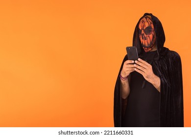 Person Wearing Jack O Lantern Pumpkin Mask And Black Hooded Cape, Consulting His Mobile Phone, Celebrating Halloween, On Orange Background. Celebration Concept, All Souls' Day And All Saints' Day.