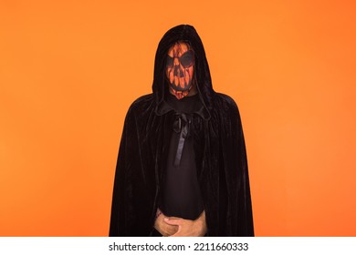 Person With Wearing Jack O Lantern Pumpkin Mask And Black Hooded Cape, Celebrating Halloween, On Orange Background. Celebration Concept, All Souls' Day And All Saints' Day.