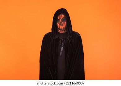 Person With Wearing Jack O Lantern Pumpkin Mask And Black Hooded Cape, Celebrating Halloween, On Orange Background. Celebration Concept, All Souls' Day And All Saints' Day.