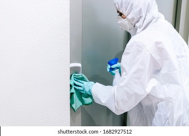 A person wearing a mask, gloves and a safety suit cleans and disinfects a doorway of a community flat in the face of a virus pandemic, protected safety equipment so as not to be infected - Shutterstock ID 1682927512