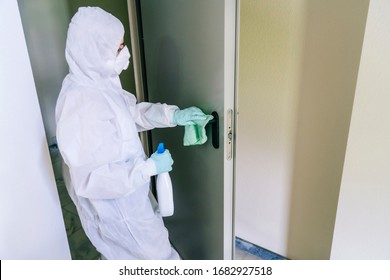A person wearing a mask, gloves and a safety suit cleans and disinfects a doorway of a community flat in the face of a virus pandemic, protected safety equipment so as not to be infected - Shutterstock ID 1682927518