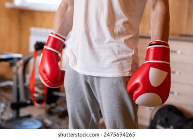 A person wearing red and white boxing gloves stands in a casual home gym setting with wooden floors and fitness equipment in the background. They are dressed in a white t-shirt and gray sweatpants