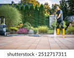 A person wearing yellow rubber boots uses a pressure washer to clean a patio area in a beautiful garden. The sun shines brightly as the landscaping thrives.