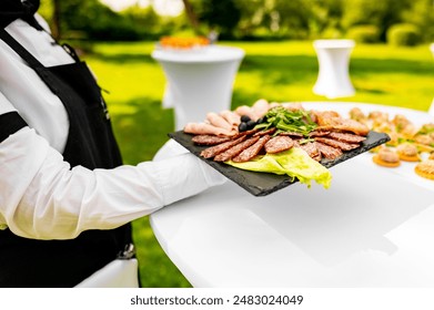 A person in a white shirt and black vest holds a tray with sliced grilled meat and greens at an outdoor catering event - Powered by Shutterstock