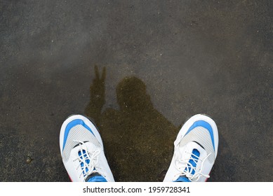 Person in white sneakers standing on the wet asphalt after the rain and taking a selfie in the reflection and puddle. Pov, top view