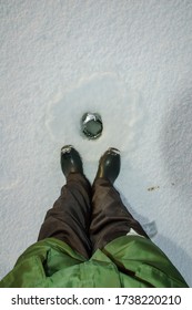 A Person In Winter Overalls And Rubber Boots Looks Down At A Hole In The Ice Made By A Fisherman.