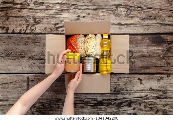 Person woman receiving donation Food box. Female\
volunteer hands packing donation box with food items of staple\
products on wooden table. Donate Food delivery concept. Donations\
grocery canned food.