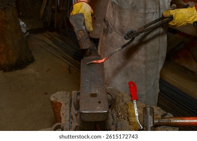 A person working hot metal on an anvil with a hammer and tongs. A red-hot iron bar, protective gloves, and traditional blacksmith tools are visible.
