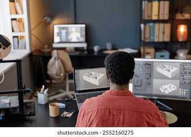 Person working on 3D design project with two computer monitors and a 3D printer in home office setup with bookshelves and work desk