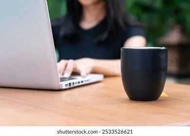 A person working on a laptop at a wooden table, with a black coffee cup in the foreground. The background features greenery, creating a relaxed outdoor workspace atmosphere. - Powered by Shutterstock