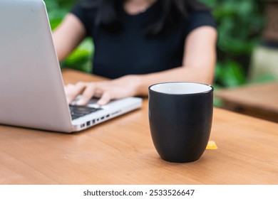 A person working on a laptop at a wooden table, with a black coffee cup in the foreground. The background features greenery, creating a relaxed outdoor workspace atmosphere. - Powered by Shutterstock