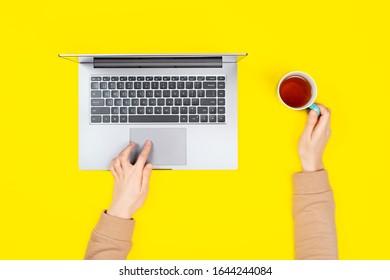 Person Working Or Typing On A Laptop Computer In A Modern Space. Female Hands Working On New Laptop On Bright Yellow Background With Tea Cup. Top View, Flat Lay.